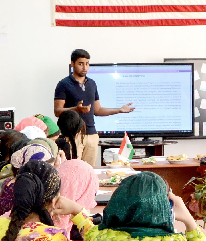 Young man stands at the front of a classroom holding hands out as he talks to women sitting in front of him most of whom are wearing head wraps