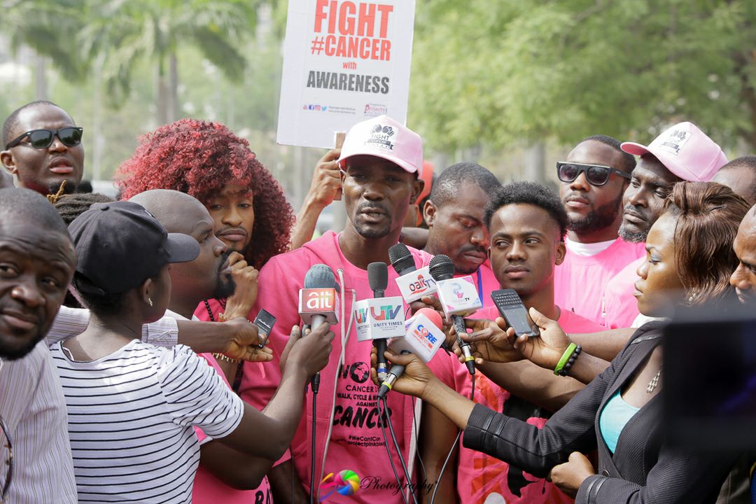 Young man stands with crowd in front of several mics and reporters appearing to interview him