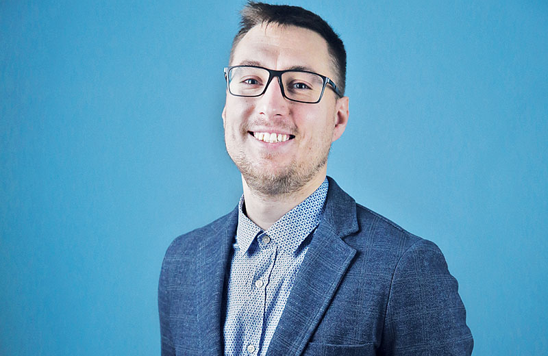 Smiling young man with glasses wearing a blue blazer over a dot-patterned collared shirt
