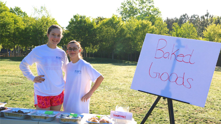 Two girls stand behind a table lined with covered plates next to a sign that says "Baked Goods"