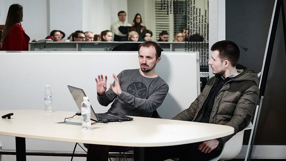 Two men talking at a table with laptop computer sitting in front of man on left