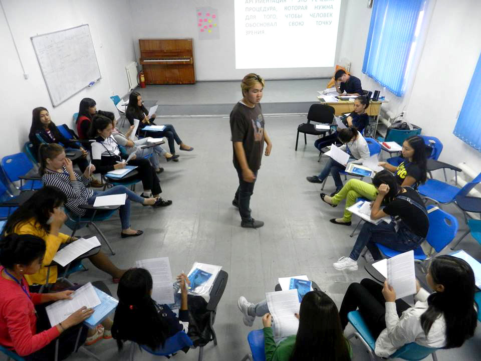 Man stands in middle of classroom surrounded by young adults sitting at desks