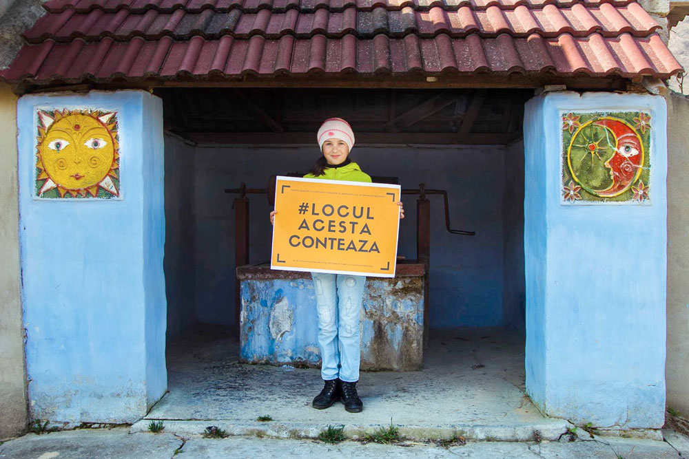 Girl stands in front of small primitive building containing well holding a sign that reads: #LOCUL ACESTA CONTEAZA