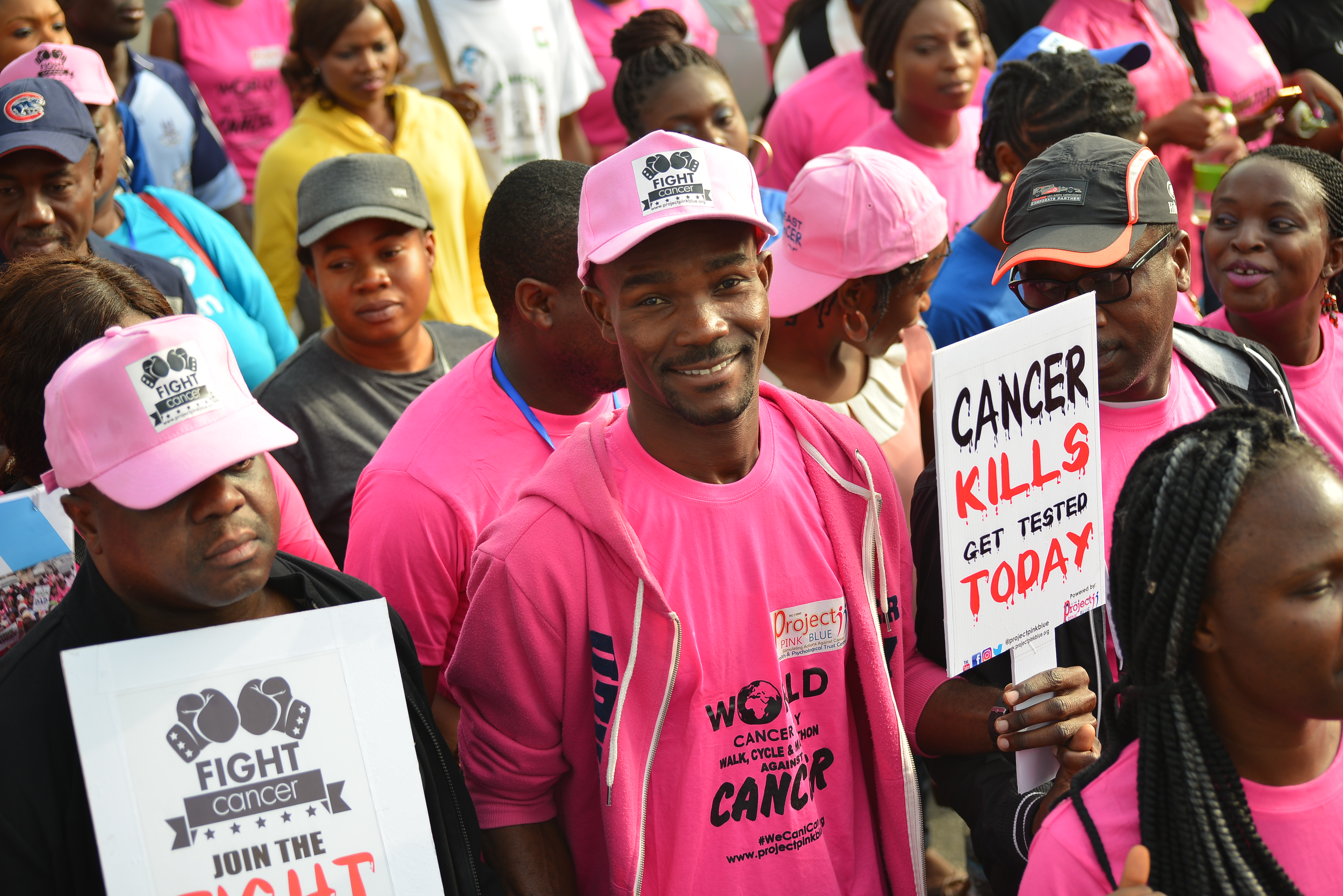 Young man stands in the middle of large crowd holding sign about Cancer