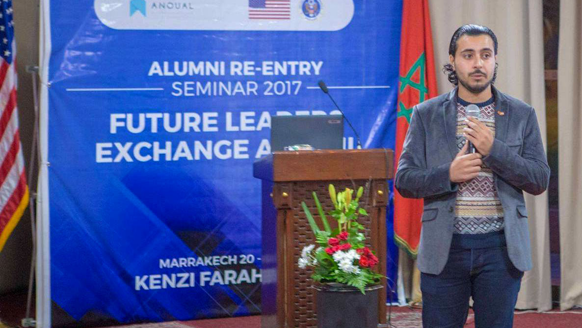 Man stands near podium holding a microphone with a large Alumni sign behind him