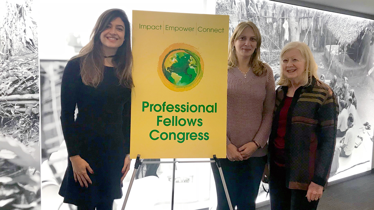 Three women stand in front of a wall lit up with an image of a rural community beside a poster that reads: Professional Fellows Congress