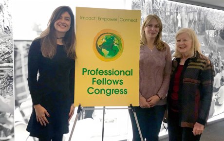 Three women stand in front of a wall lit up with an image of a rural community beside a poster that reads: Professional Fellows Congress